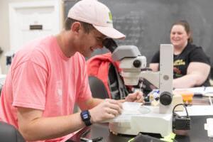 Lab technician Sara Harper-Roche laughs in the background while Ethan Caldwell looks into a microscope in the lab. 