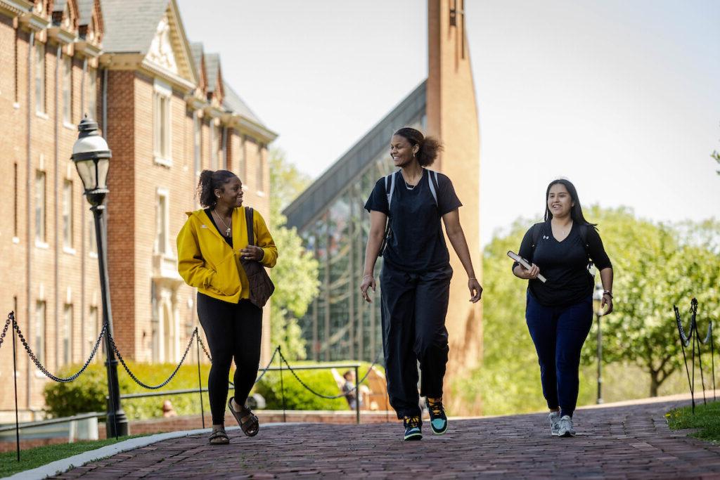 Three students walk on back campus, with the chapel in the distance behind them